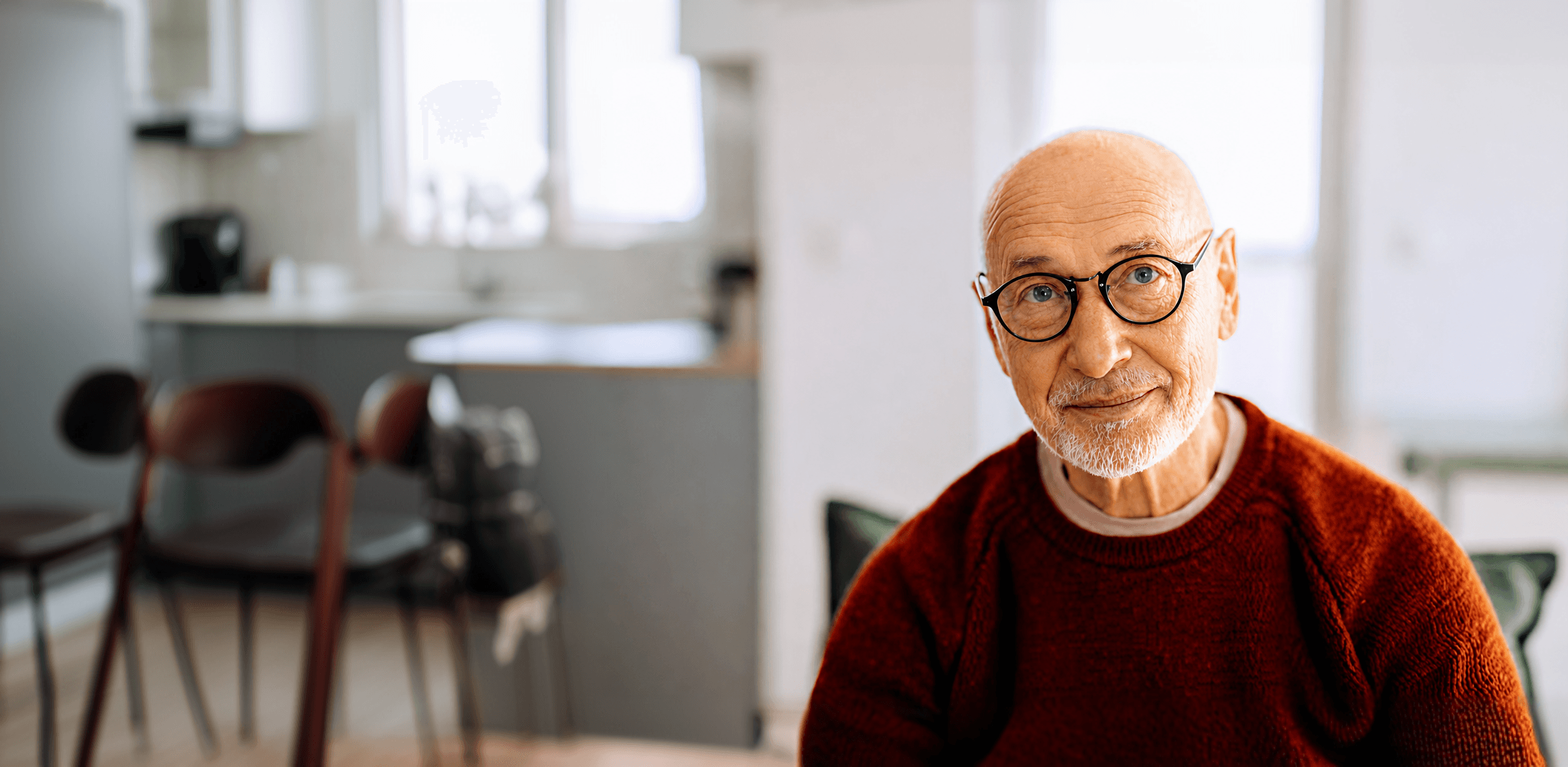full-bleed photo of an older bald man with round black glasses and a white beard, wearing a rust-red sweater, smiling softly in a bright kitchen