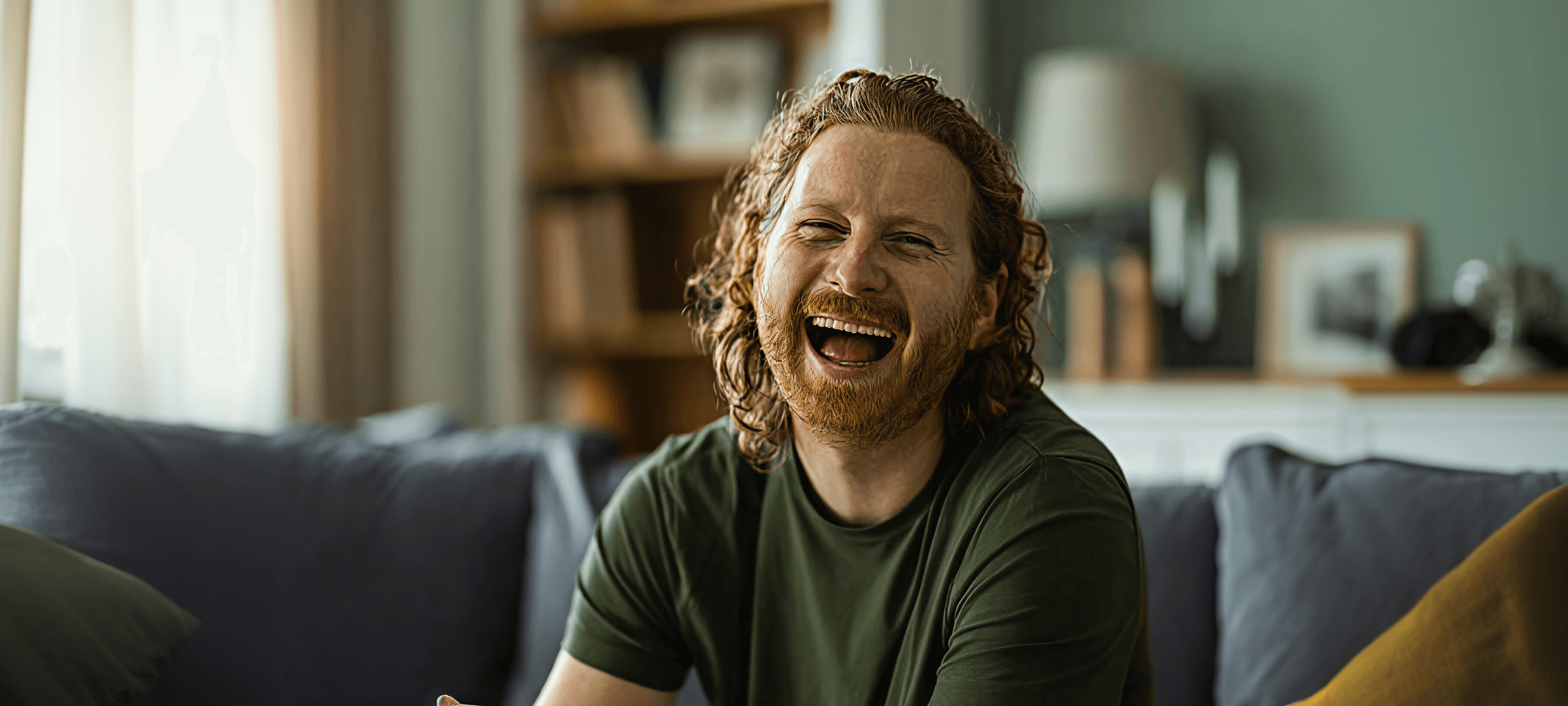 laughing bearded man with curly auburn hair on a couch in a living room